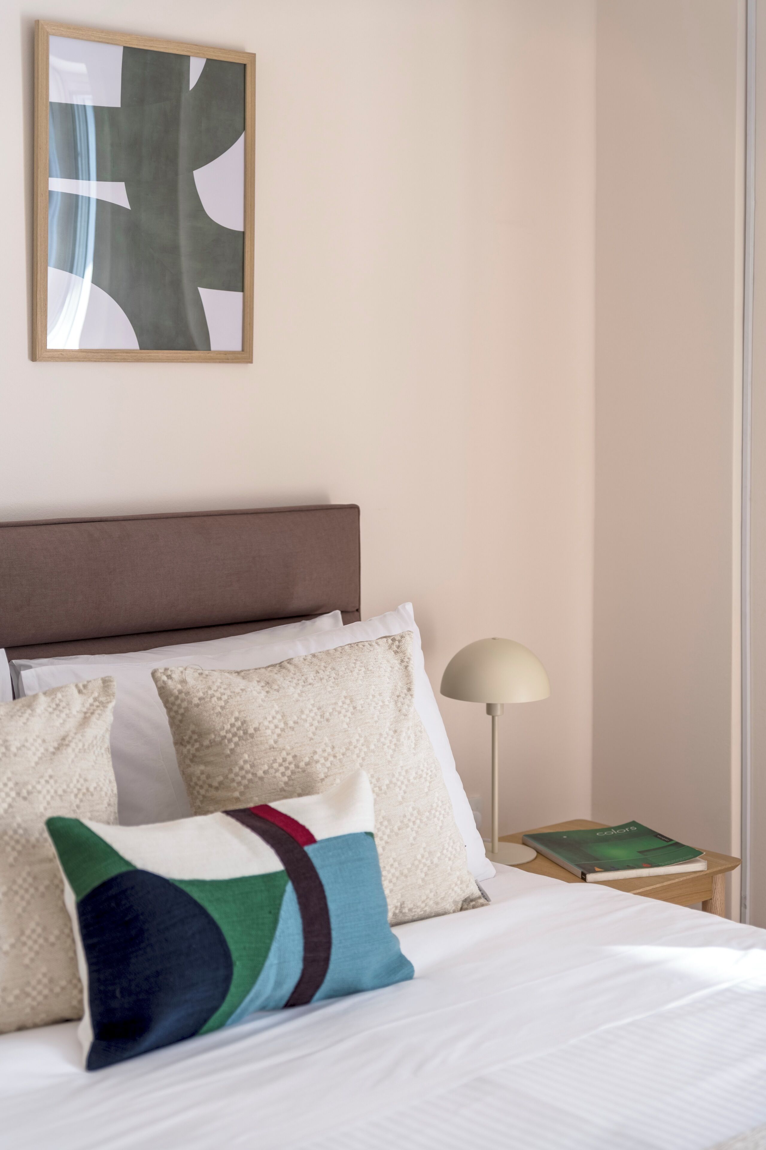 Un lit bien fait, avec du linge de lit blanc et des coussins colorés, une lampe de chevet et une œuvre d’art abstraite encadrée accrochée au mur.