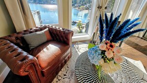 Un divano in pelle marrone con un cuscino decorato, un tavolino con un composizione floreale, e vista su un lago e edifici attraverso ampie finestre.