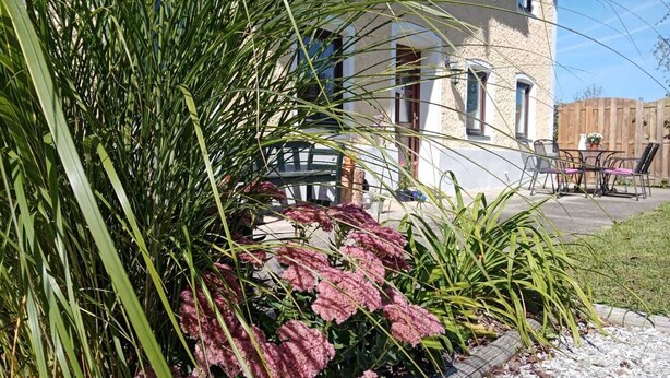Un edificio con fachada de piedra, un patio con mobiliario exterior y un jardín con diversas plantas.