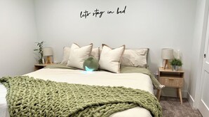 Ein ordentlich gemachtes Bett mit einer grünen Strickdecke, eine Wand mit dem Schriftzug 'Let's stay in bed' und ein Nachttisch mit einer Lampe und einer kleinen Pflanze.
