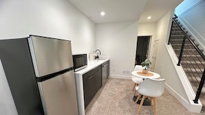 Eine kompakte Küche mit Kühlschrank, Mikrowelle und Spüle. Es gibt einen Essbereich mit Tisch und Stühlen, und eine Treppe, die zu einer weiteren Ebene führt.