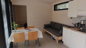A compact living space with a dining table, chairs, a sofa, a small kitchen area with a sink and coffee maker, and a window with a view of trees. - Pistohiekka Resort (Puumala)