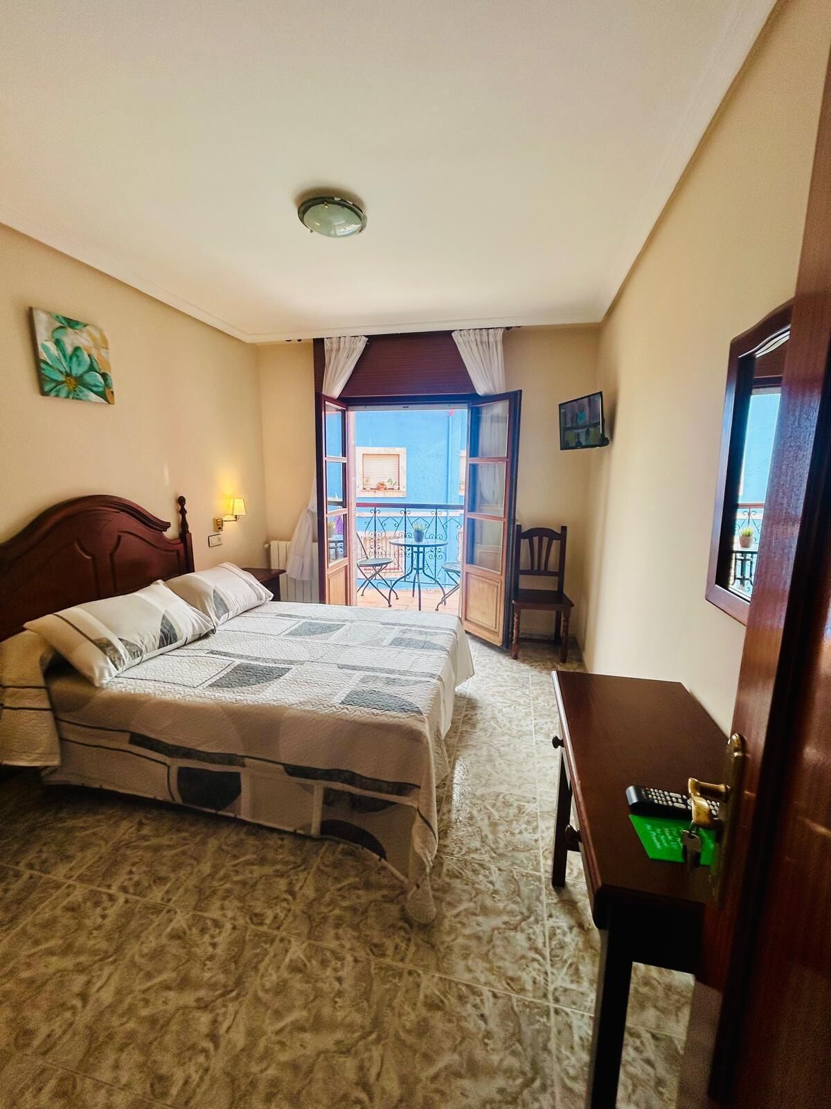A bedroom with a bed, a desk, a chair, a balcony with a table and chairs, and a view of the outside.
