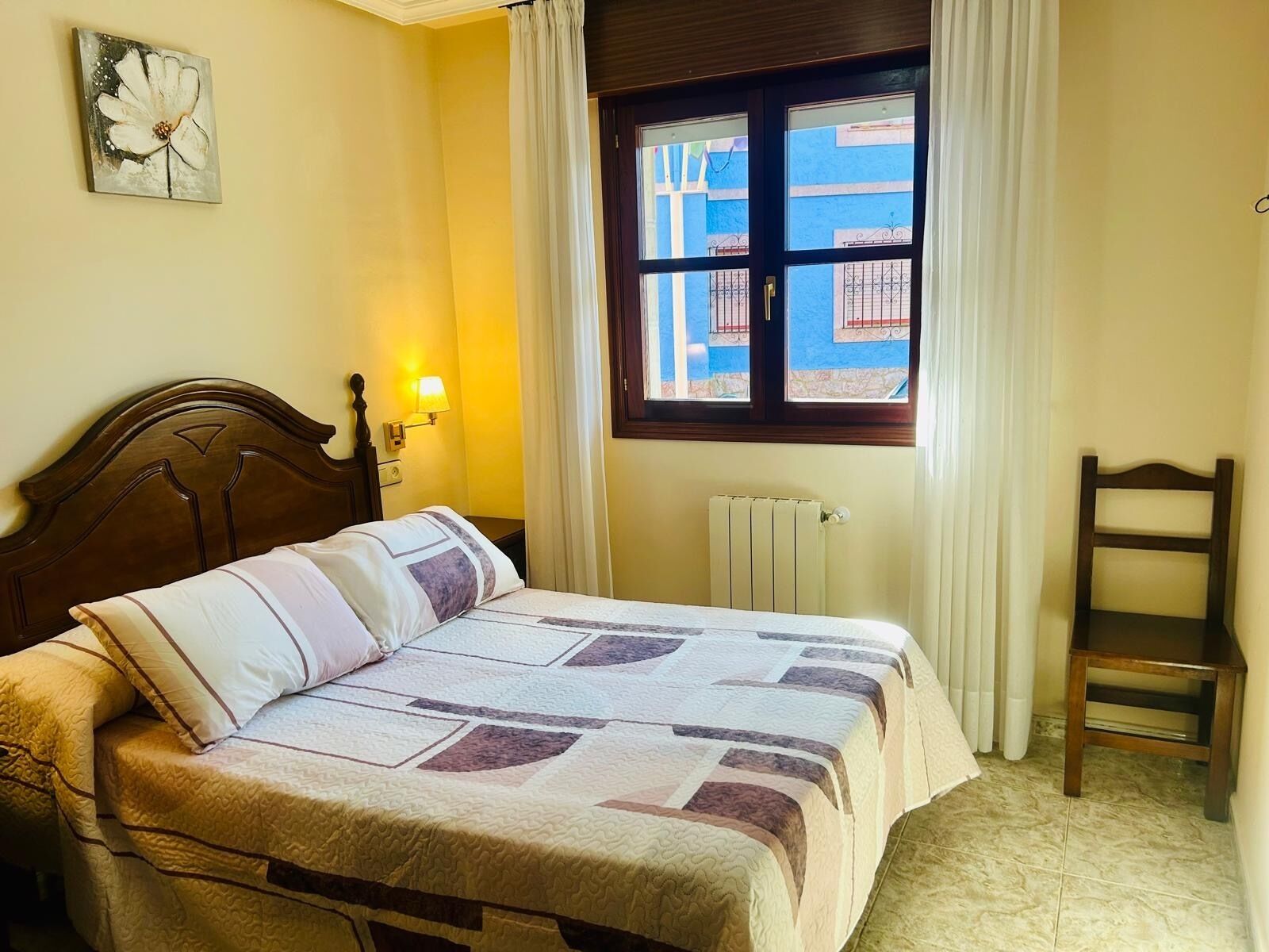 A neatly made bed with a patterned bedspread, a wooden headboard, a bedside lamp, a window with curtains, and a radiator.