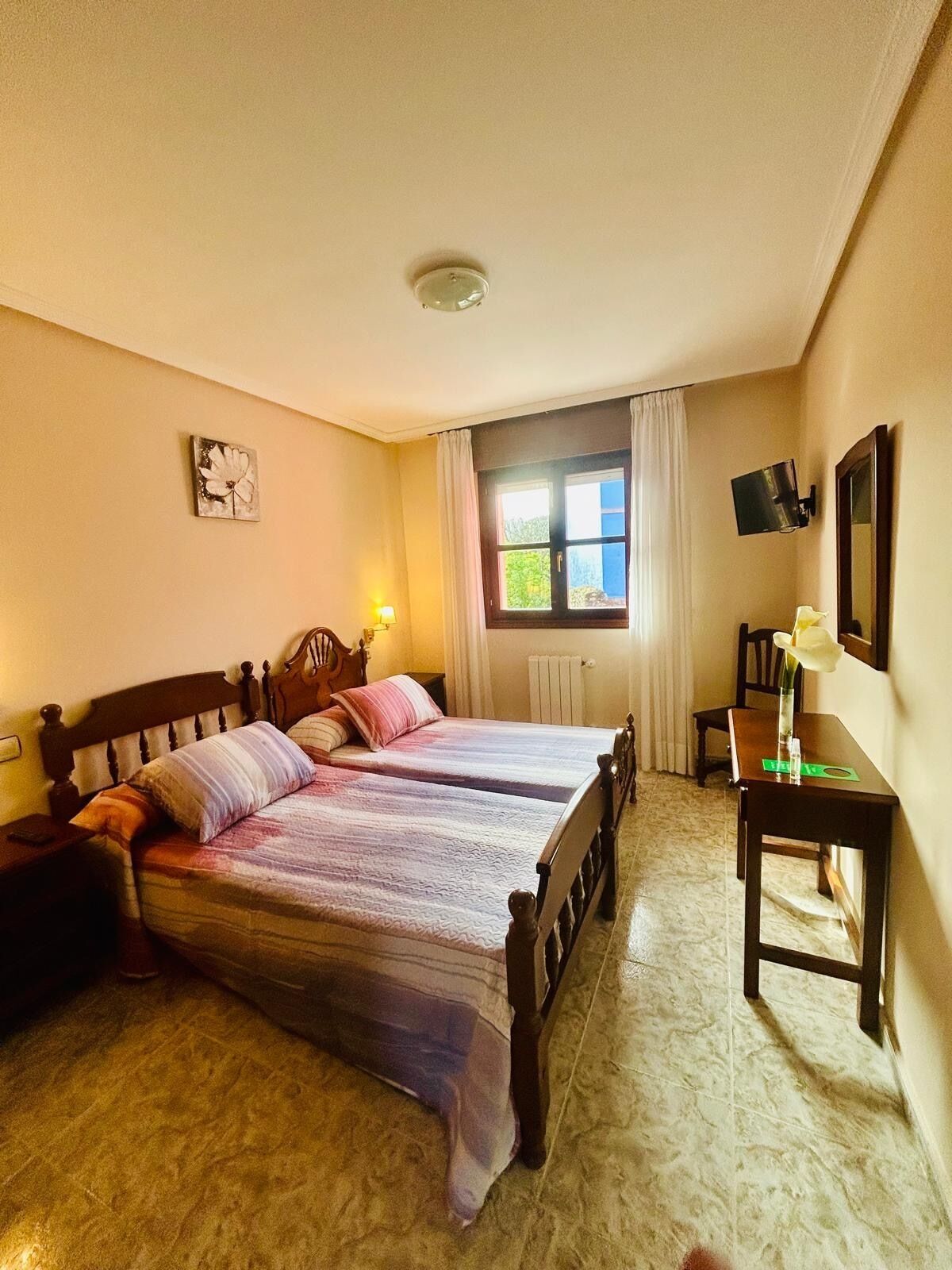 A bedroom with two beds, a desk, a chair, a window with curtains, a radiator, and a wall-mounted TV.