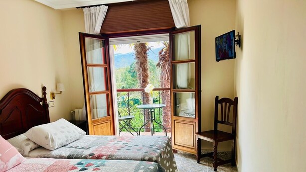 A bedroom with two beds, a small table, a chair, and a balcony with a view of greenery. - Hotel Las Palmeras (Celorio)