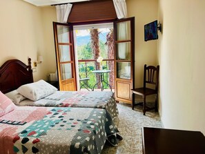 Ein Schlafzimmer mit zwei Betten, einem kleinen Tisch, einem Stuhl und einem Balkon mit Blick ins Grüne.