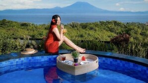 A person relaxing by a pool with a tray of items, overlooking a landscape with a mountain and the sea. - Reynten Hill  (Penida Island)