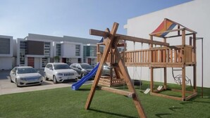 Une structure de jeu en bois, dotée d’un toboggan, se dresse devant un bâtiment moderne, avec plusieurs voitures garées dans l’allée.