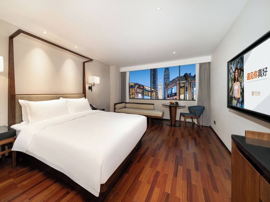 A hotel room with a large bed, a desk, a chair, a small table, and a sofa. There is a window with a cityscape view.