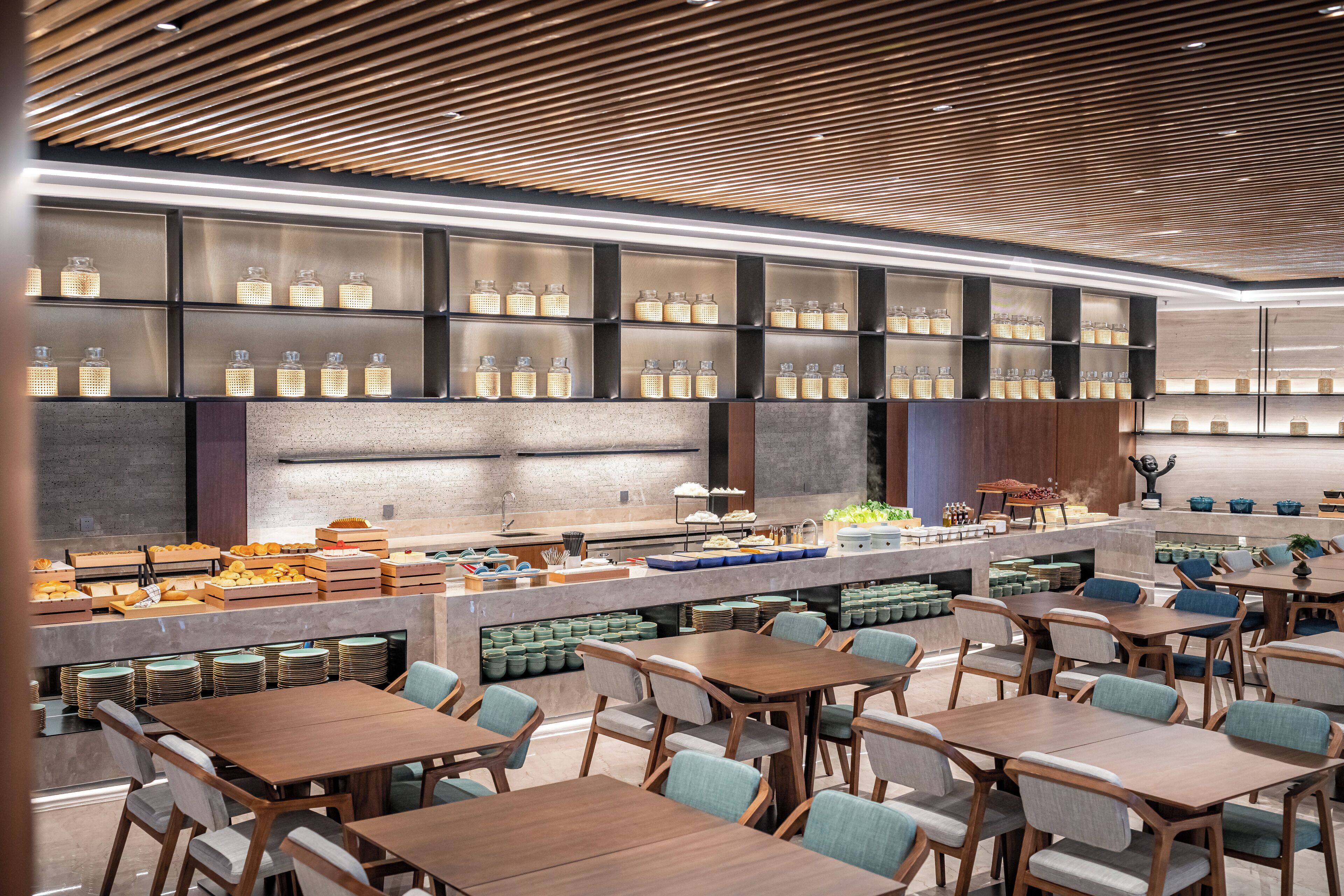 A modern restaurant with wooden tables and chairs, a counter with food items, and shelves with bottles.