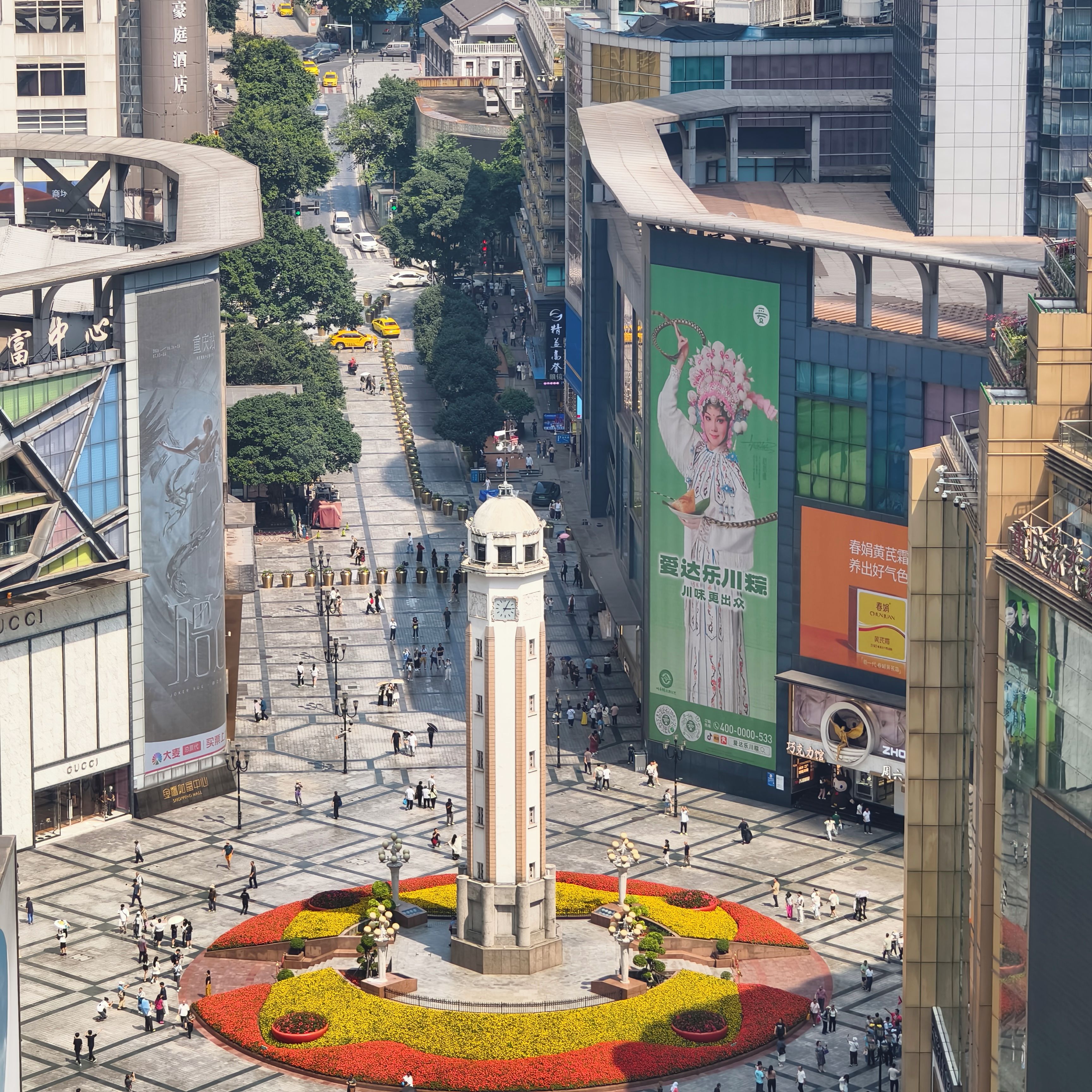 Une place publique avec un monument central, entourée de parterres de fleurs et d’immeubles modernes.