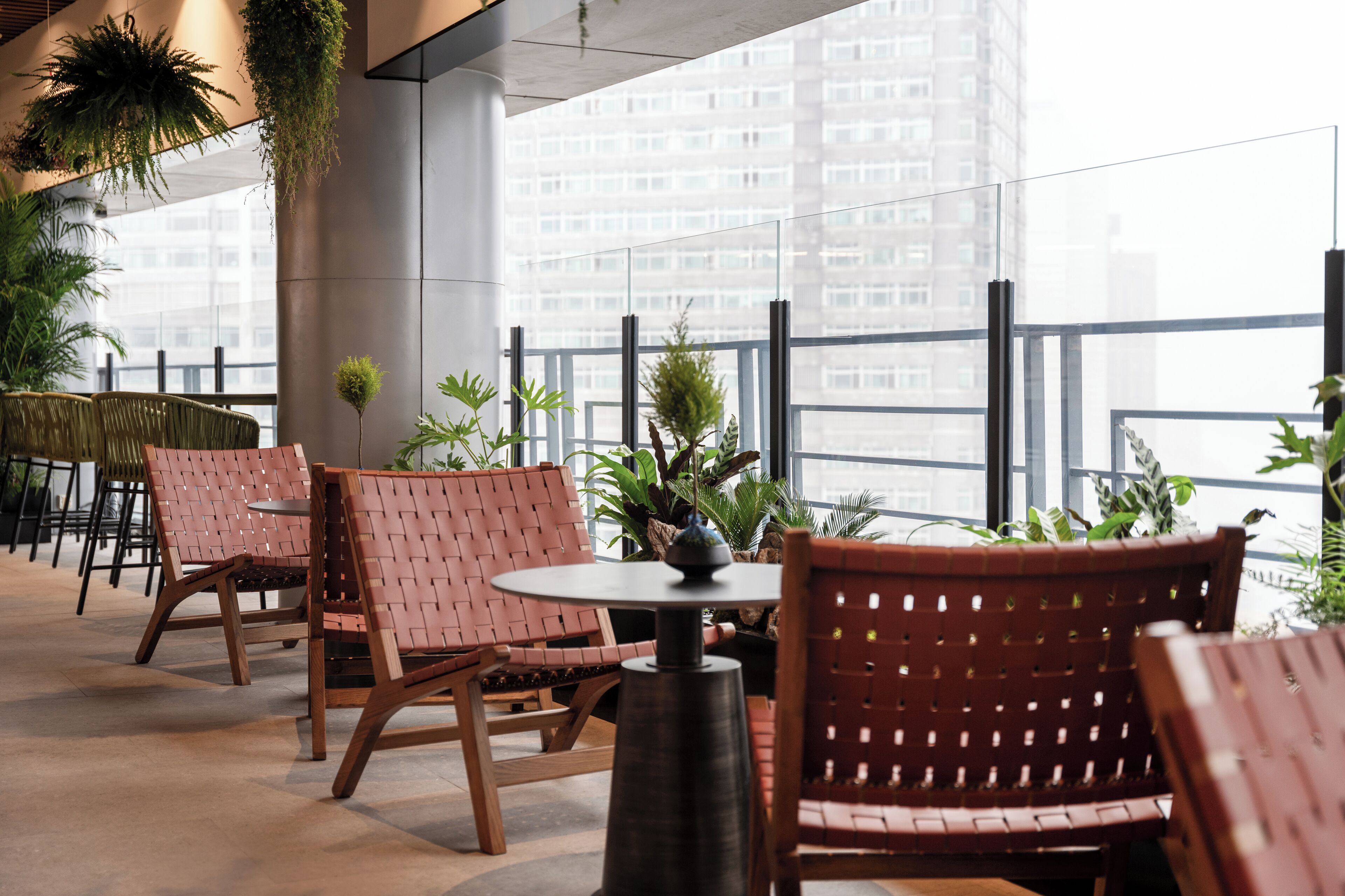 Een moderne buitenruimte met rieten stoelen en tafels, omgeven door potplanten en met uitzicht op een stadsbeeld.