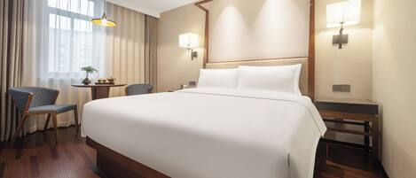 Habitación de hotel con una cama grande, un escritorio con silla, una lámpara y un ventanal con cortinas.