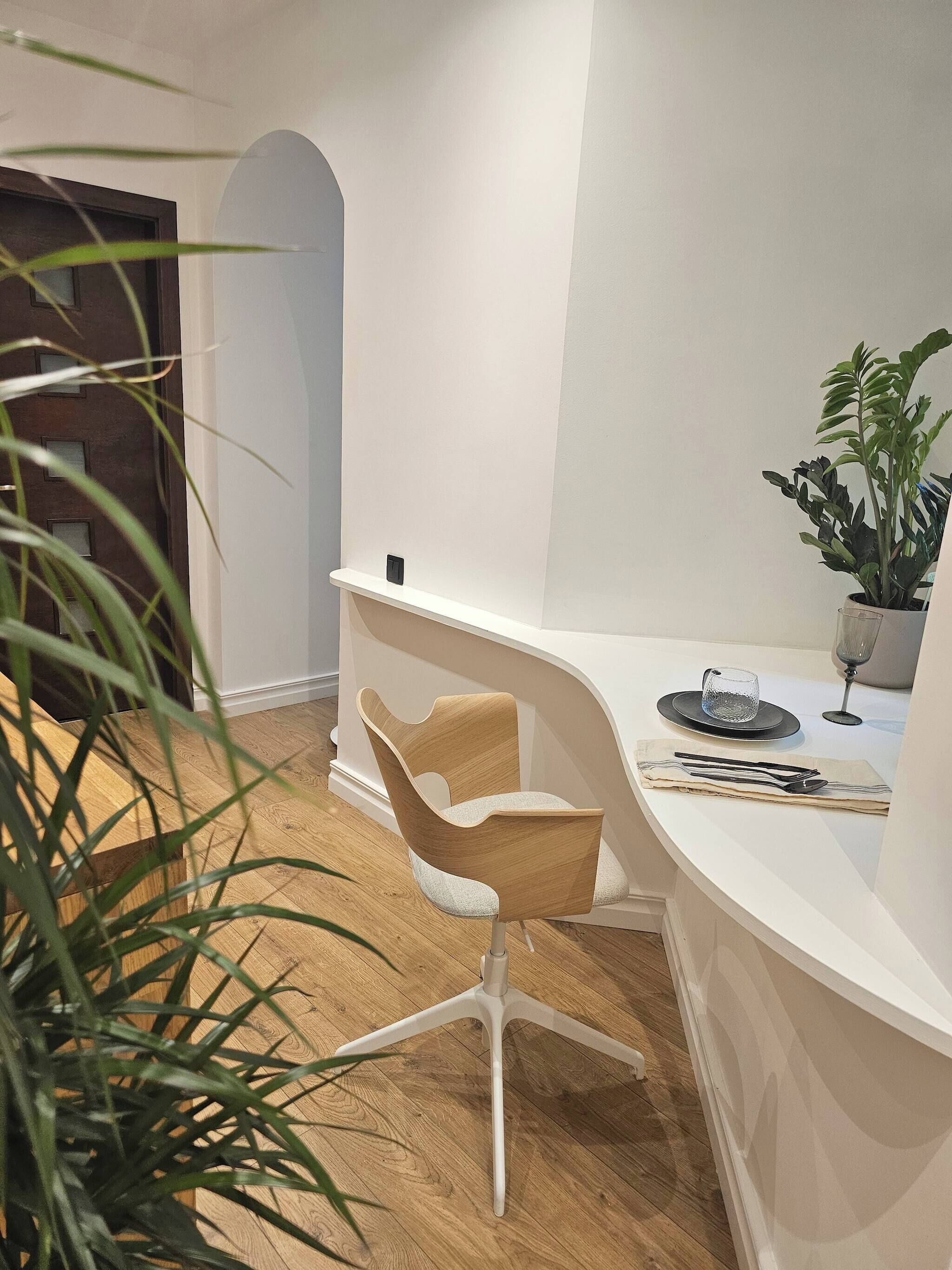 A modern, minimalist room with a curved white reception desk, a wooden chair, a potted plant, and a glass of water on the desk.