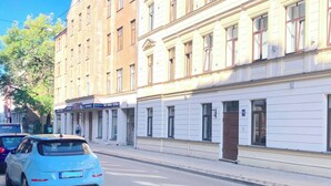Une rue bordée d’immeubles à plusieurs étages, une voiture bleue garée sur le bord et un ciel bleu dégagé.