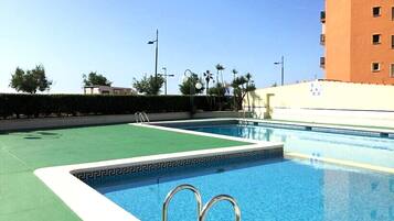 Una piscina con cubierta verde y borde blanco, rodeada de seto y palmeras.