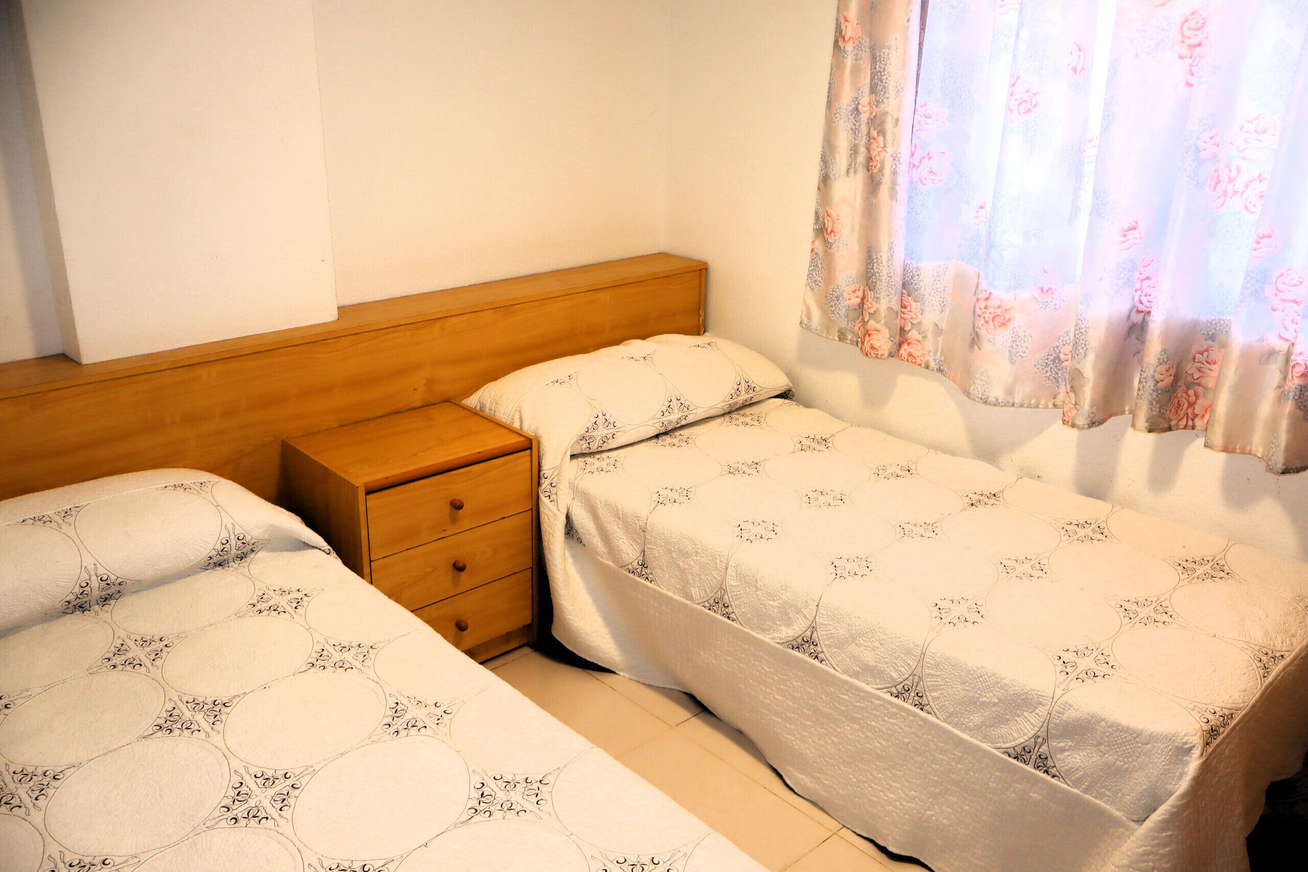 A bedroom with two single beds, a wooden nightstand, and a window with floral curtains.
