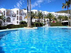 A swimming pool with clear blue water, surrounded by white buildings with balconies and palm trees. - Calas atico con aire y barbacoa (Peñiscola)