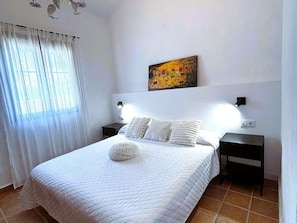 Een slaapkamer met een bed, witte gordijnen, een schilderij, twee bedlampen en een stopcontact.
