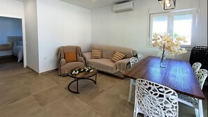 Una sala moderna con un sofá color beige, una mesa de comedor de madera y sillas blancas. Hay una pequeña mesa redonda con un centro de mesa floral. La habitación tiene un ventanal con cortinas blancas y una lámpara de techo.