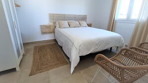 Un dormitorio con cama, cabecera de madera, alfombra tejida, sillas de mimbre y una ventana con cortinas.