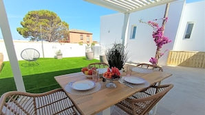 Un patio con una mesa de madera dispuesta para cuatro personas, rodeado de sillas de mimbre, y una pérgola blanca encima.