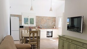 A modern kitchen with white cabinets, a built-in oven, and a microwave. There is a dining area with a wooden table and chairs, and a green cabinet with drawers. A flat-screen TV is mounted on the wall. - Suite La Morera in Conil de la Frontera (Conil de la Frontera)