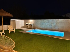 A backyard with a swimming pool, a hot tub, a thatched umbrella, and a seating area with chairs.