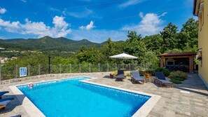 Une piscine rectangulaire, dotée de chaises longues et d’une pergola, entourée d’une terrasse en pierre et offrant une vue sur les montagnes.
