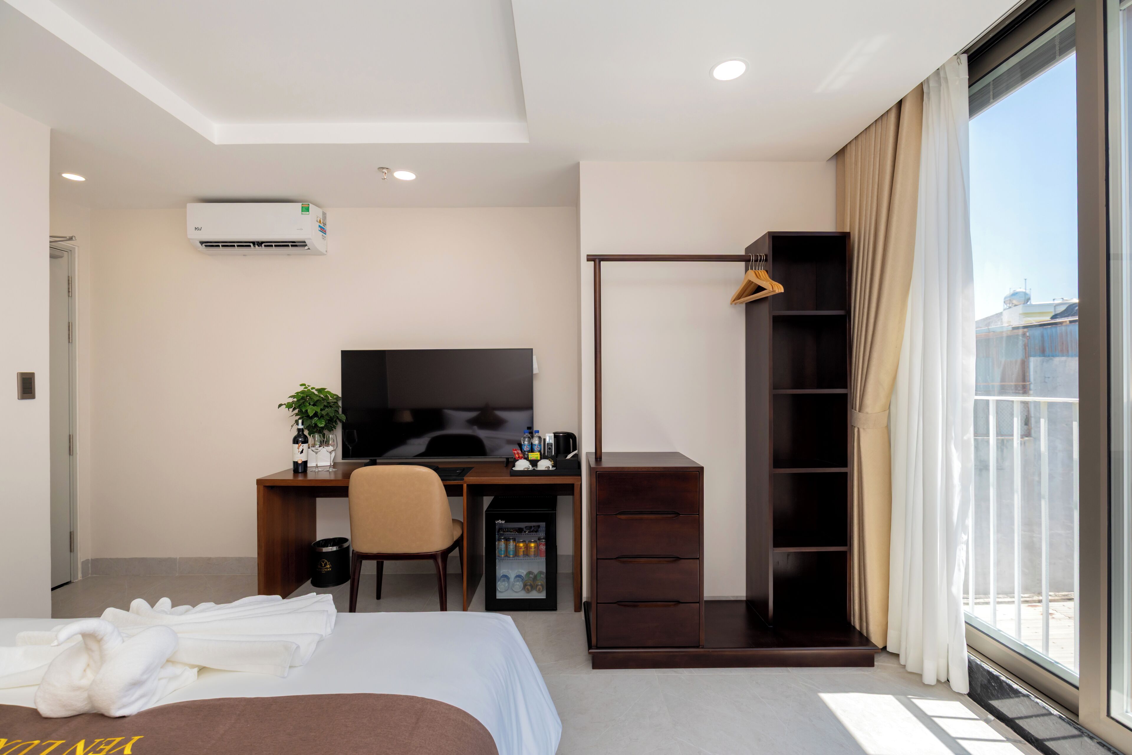 A modern hotel room with a bed, a desk with a computer, a TV, a mini-fridge, and a wardrobe.