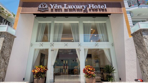 The entrance of the Yen Luxury Hotel with a modern facade, large glass doors, and two large flower pots in front.