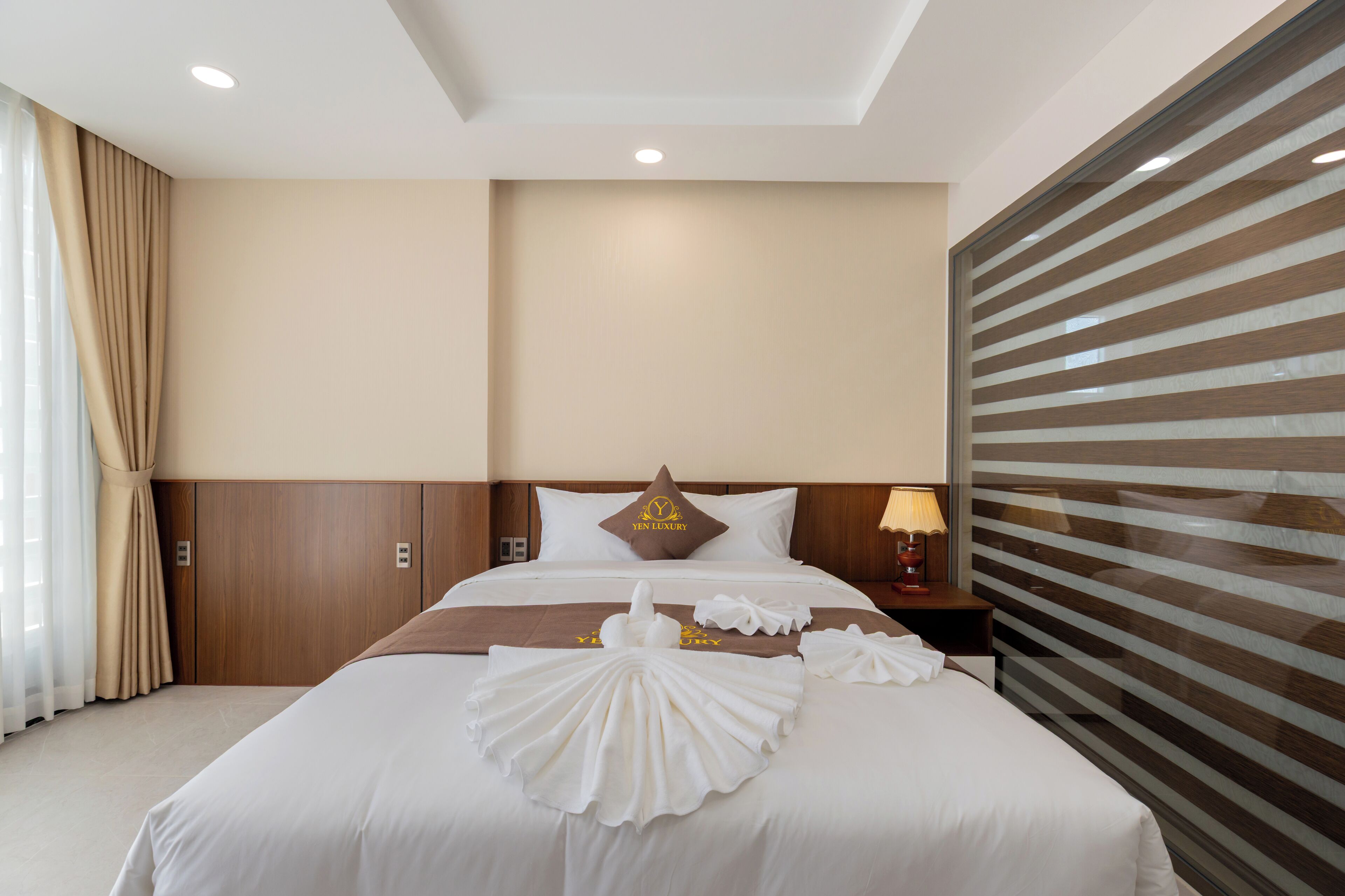 A hotel room with a neatly made bed, a headboard with a logo, a nightstand with a lamp, and a large window with blinds.