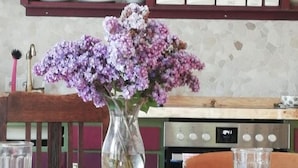 A dining area with a wooden table, two chairs, a vase with purple flowers, and a kitchen in the background. - Vacation apartment Am Schloßberg (Solingen)