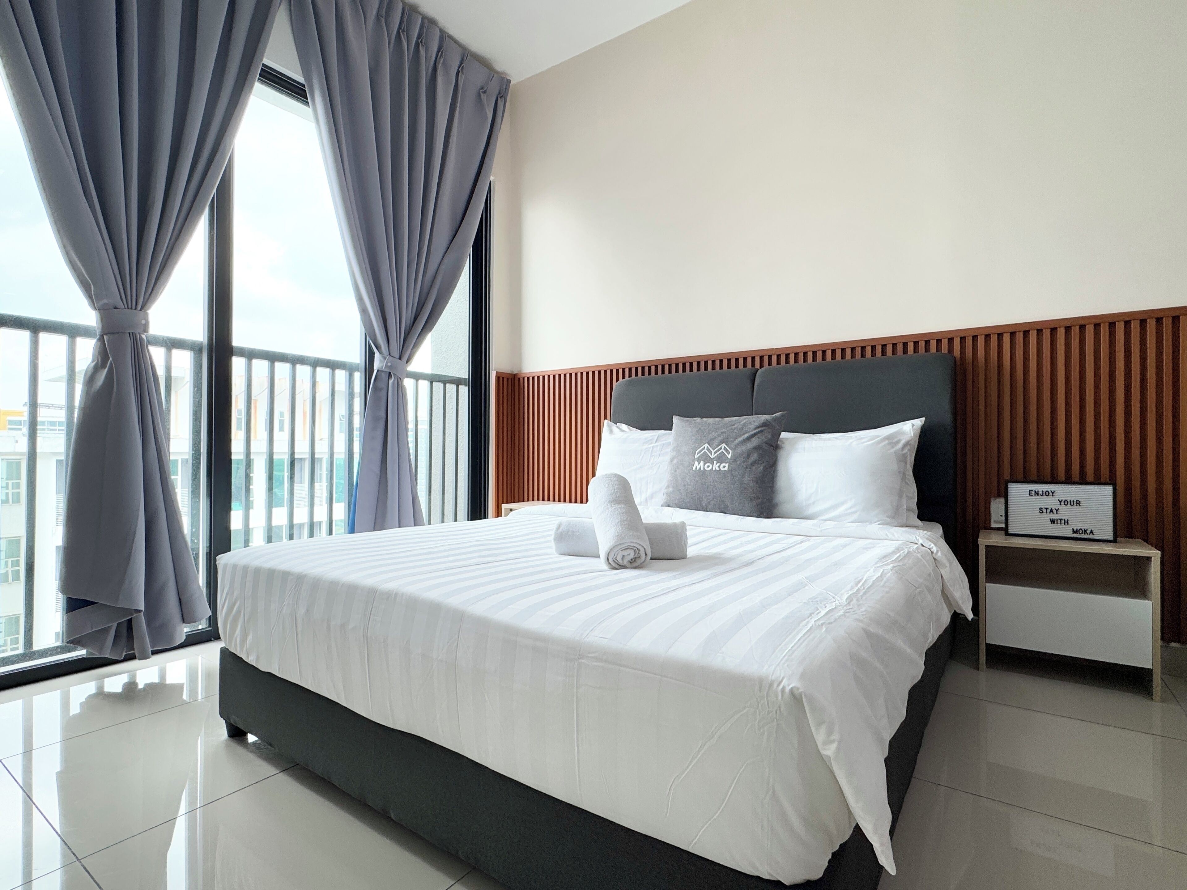 Une chambre d’hôtel moderne avec un grand lit, des rideaux gris, une tête de lit en bois et une table de chevet sur laquelle est posé un panneau.