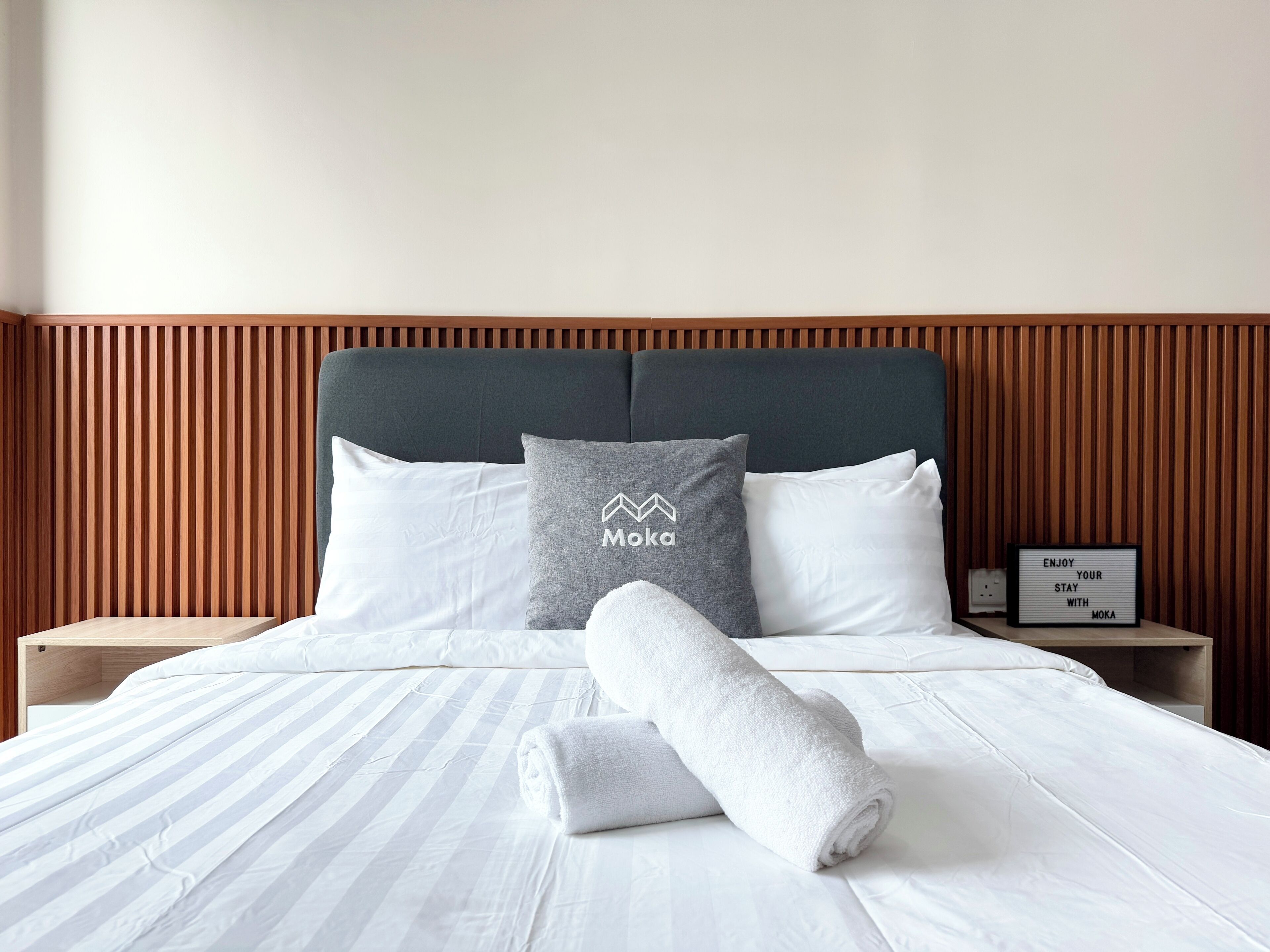 Een net opgemaakt bed met wit beddengoed en een grijs kussen met een logo, een opgerolde witte handdoek en een klein bordje op het nachtkastje.