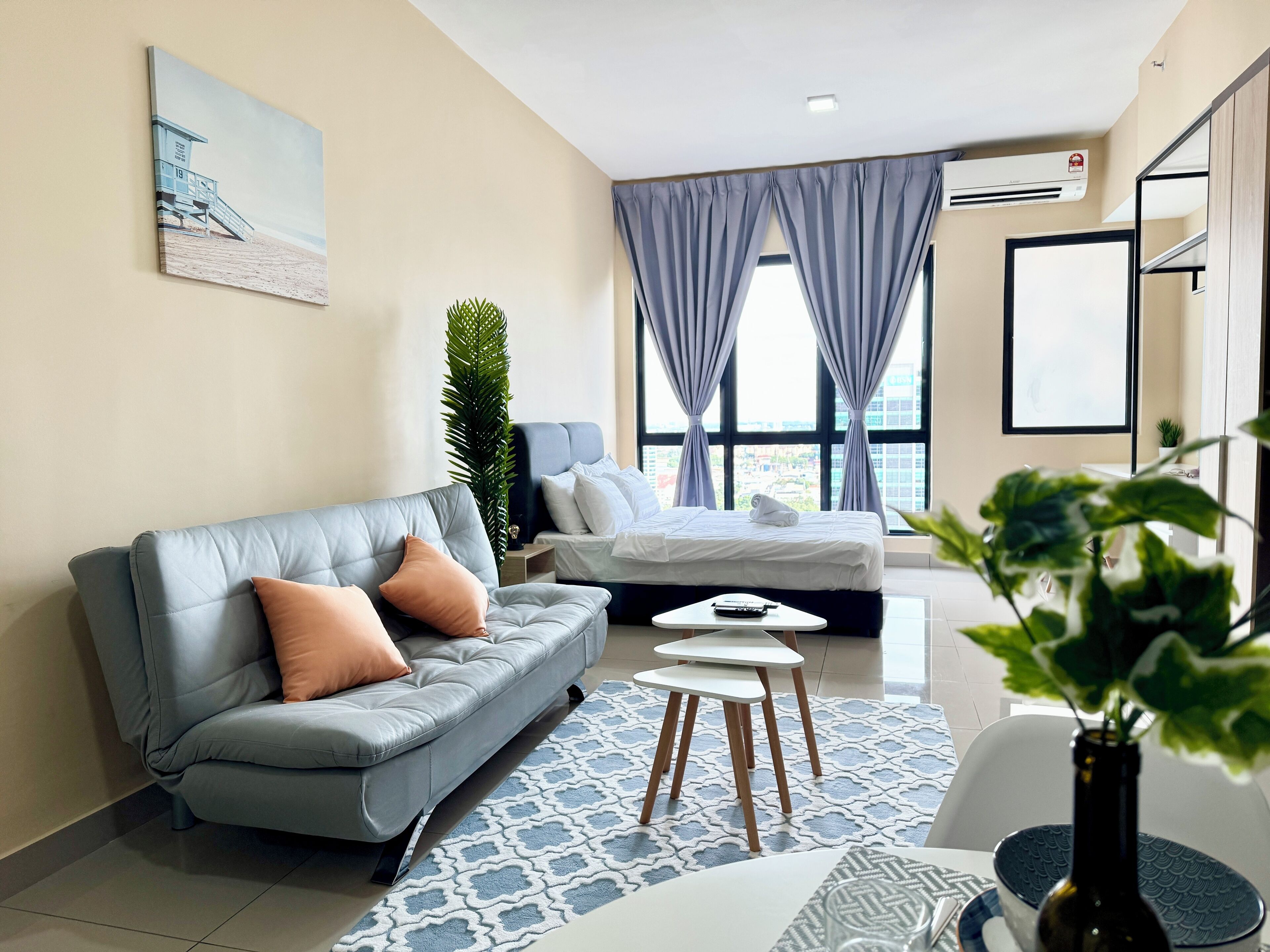 Habitación de hotel moderna con sofá gris, cama, mesita y un amplio ventanal con cortinas.