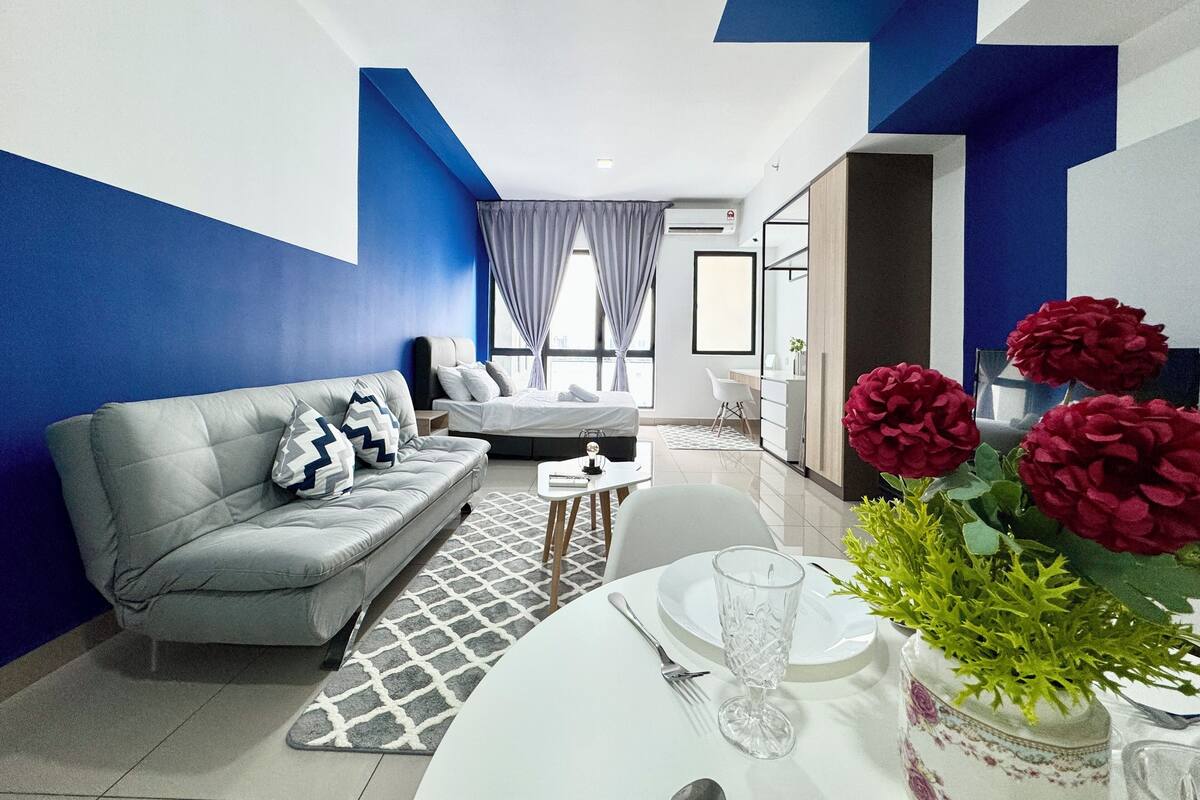 A modern hotel room with a grey sofa, a dining table set for two, and a large window with curtains.
