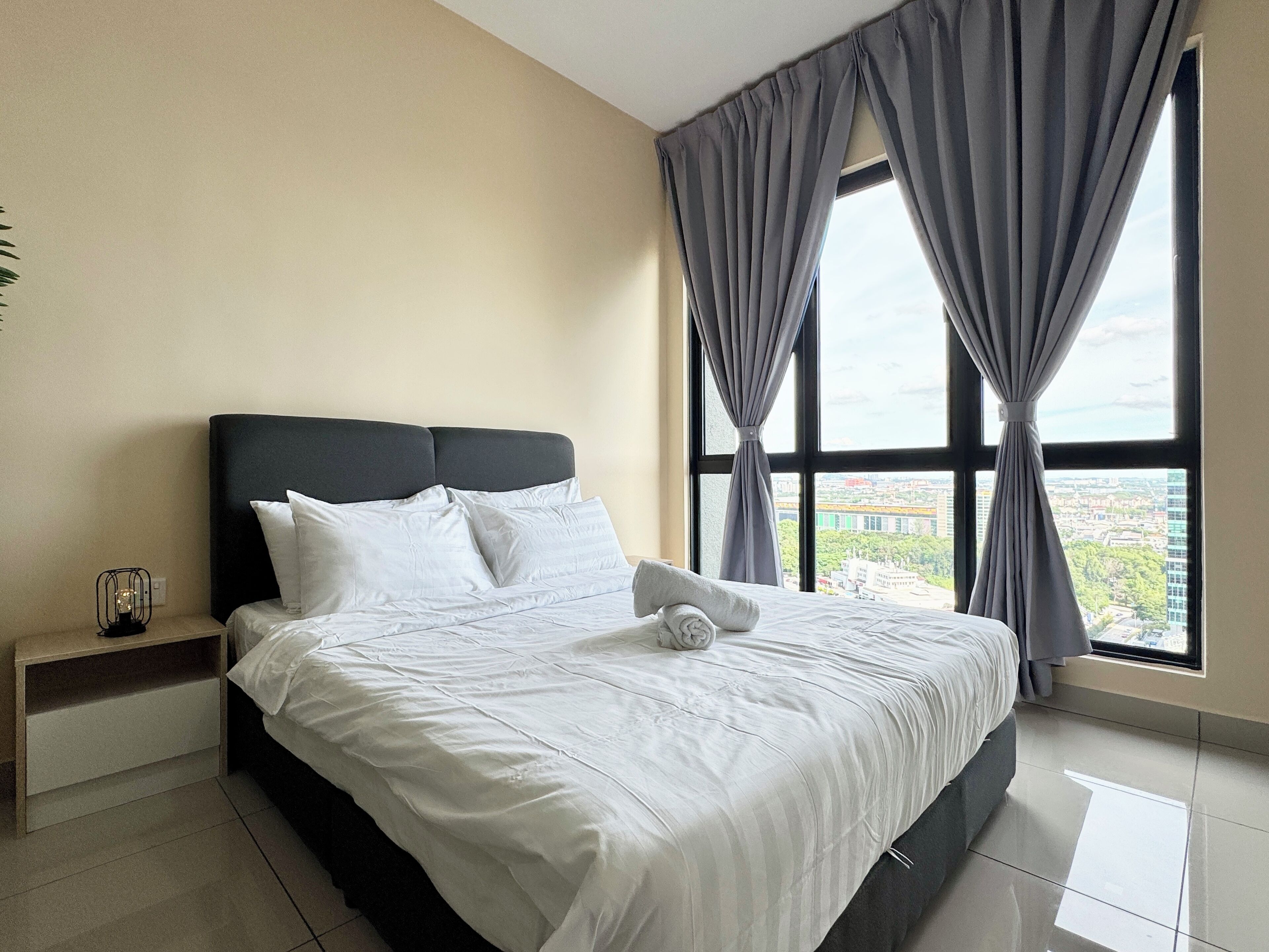 Una cama bien hecha con sábanas y almohadas blancas, una mesita de noche con lámpara, y vistas a un paisaje urbano a través de amplios ventanales con cortinas grises.
