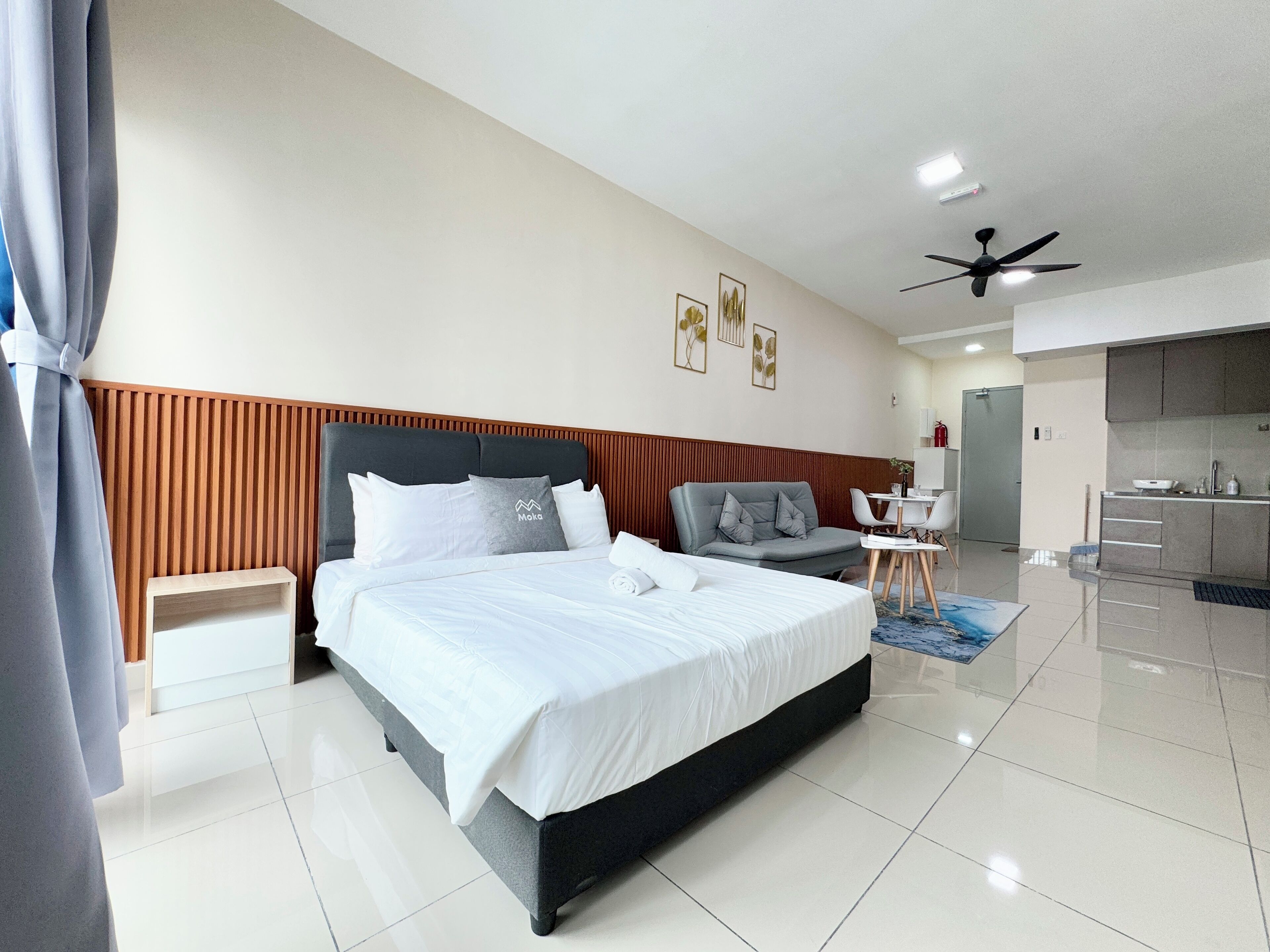 Une chambre d’hôtel moderne avec un grand lit, un canapé gris, une petite table à manger et un coin cuisine.
