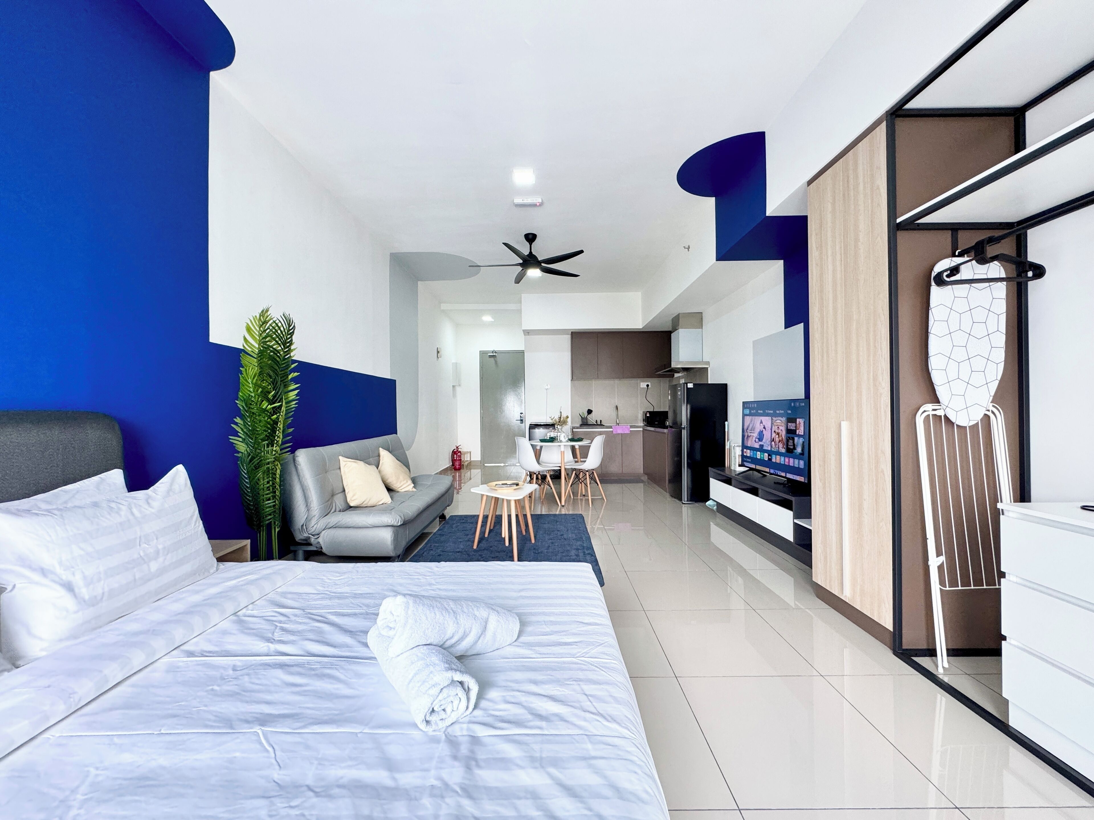 A modern hotel room with a large bed, a seating area with a sofa and chairs, a kitchenette, and a television.