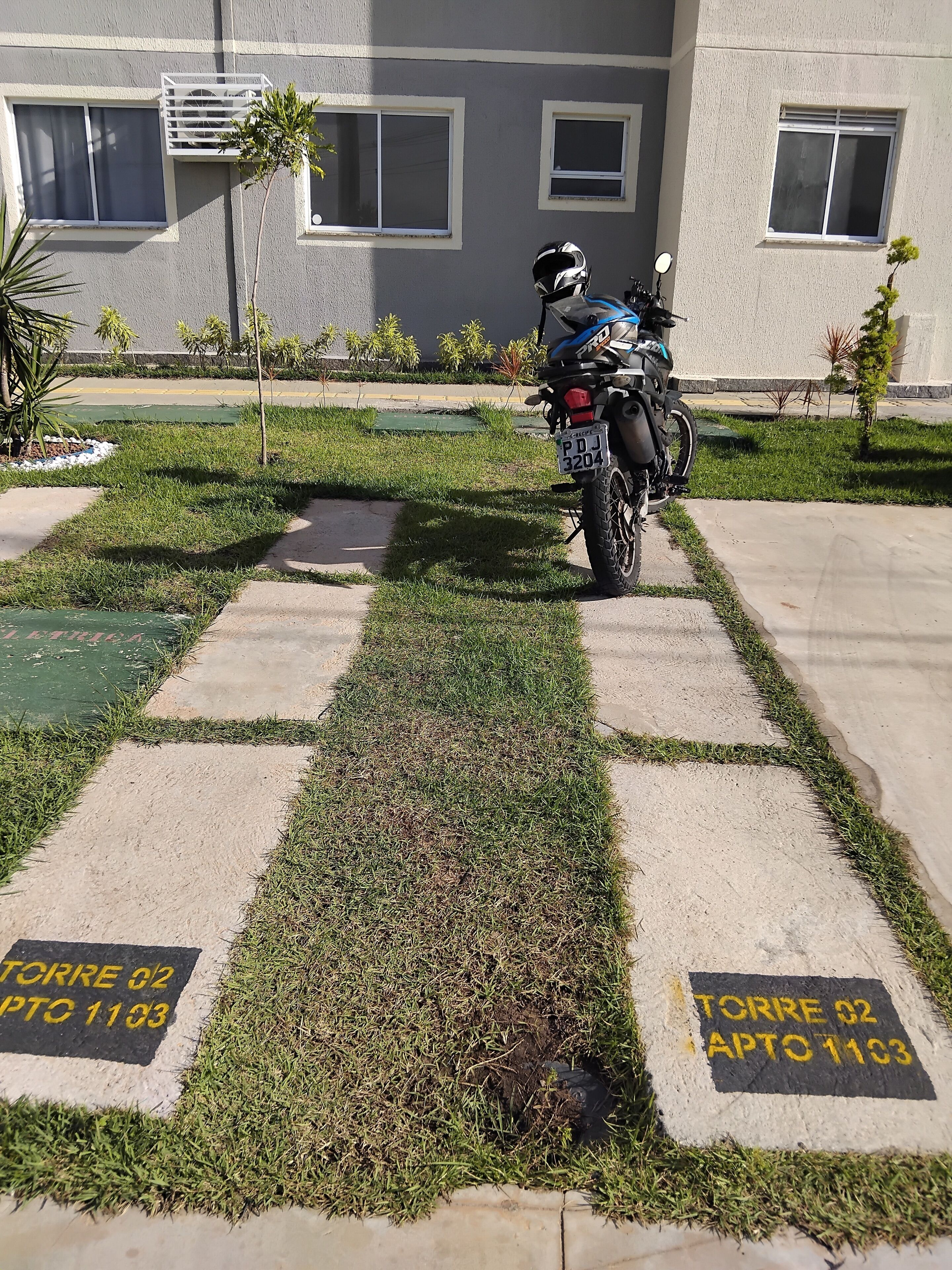 Une moto est garée sur un chemin de béton bordé d’herbe, devant un immeuble doté de fenêtres et d’une antenne parabolique.