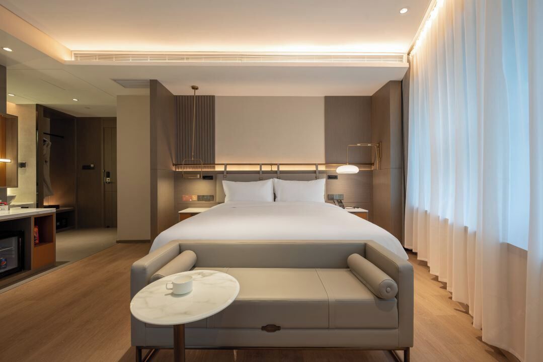 Une chambre d’hôtel moderne dotée d’un grand lit, d’un coin salon avec une table et offrant une vue sur la ville grâce à de grandes fenêtres.