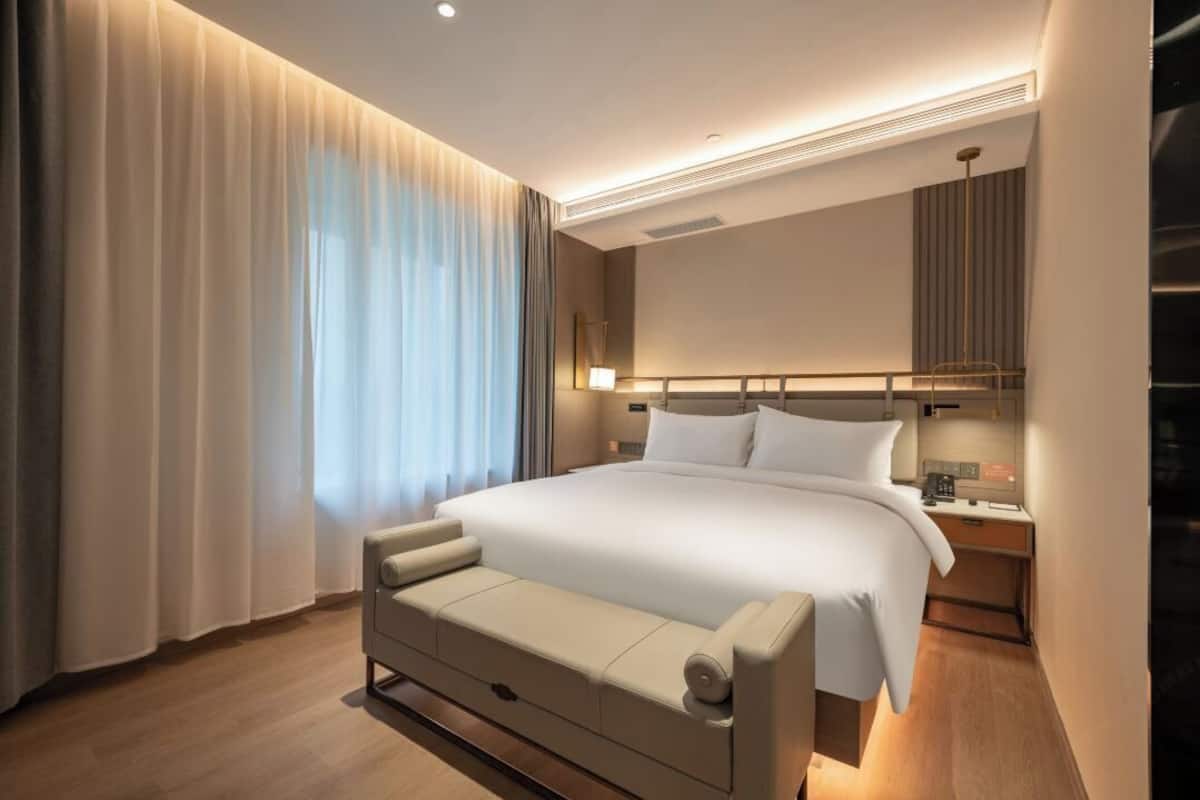 A modern hotel room with a large bed, a bedside table, a lamp, and a view of the city through the window.