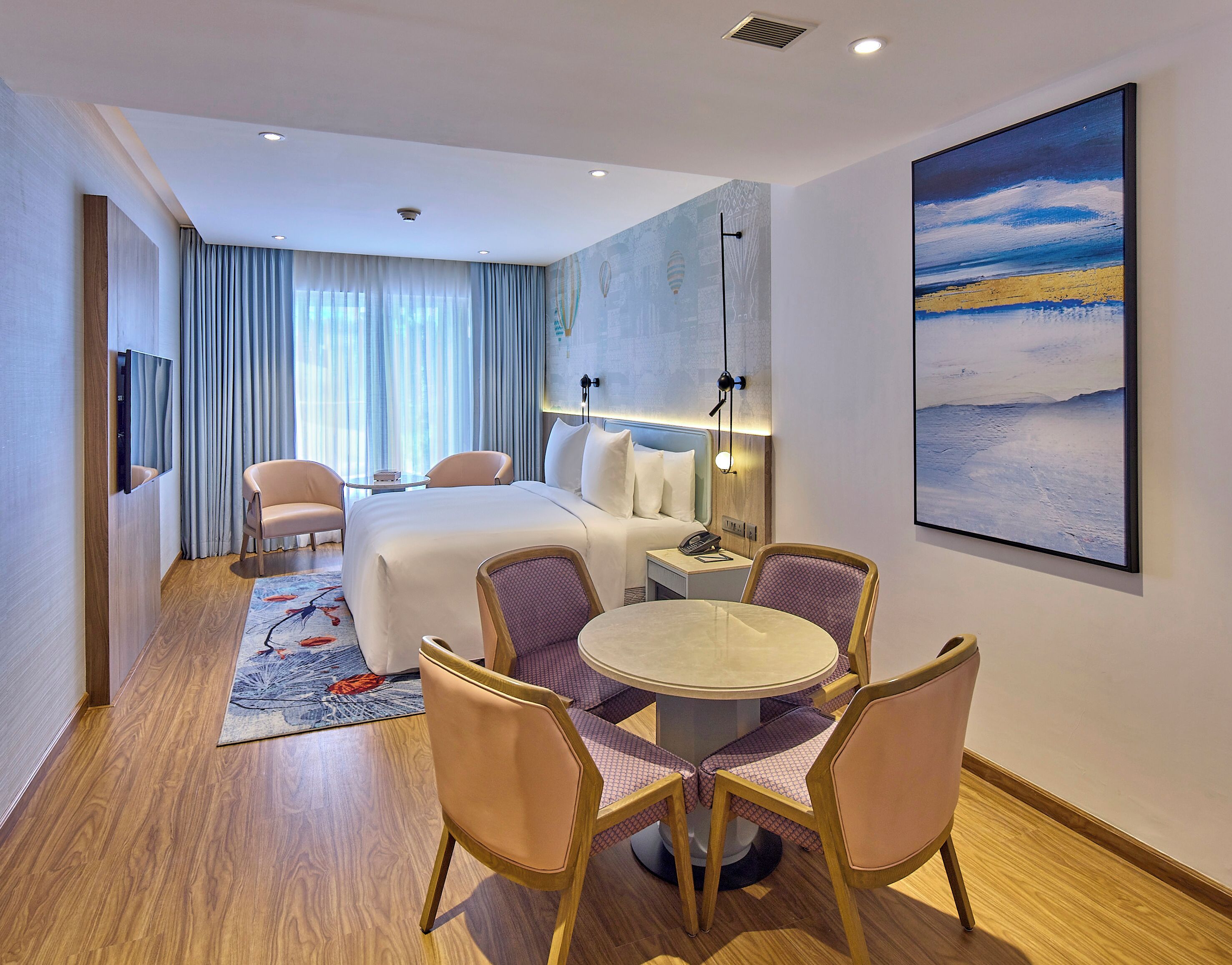 Una habitación de hotel con cama, dos sillones, una mesa de comedor con sillas, televisor, mesita de noche con lámpara y un cuadro grande en la pared.