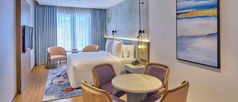 Una habitación de hotel con cama, dos sillones, una mesa de comedor con sillas, televisor, mesita de noche con lámpara y un cuadro grande en la pared.