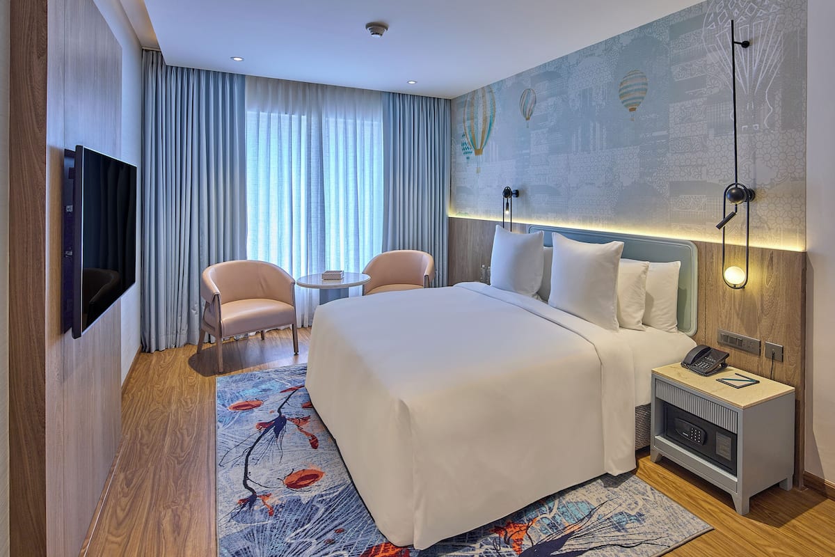 A modern hotel room with a large bed, a flat-screen TV, a small table, two chairs, a nightstand with a phone, and a decorative wall.
