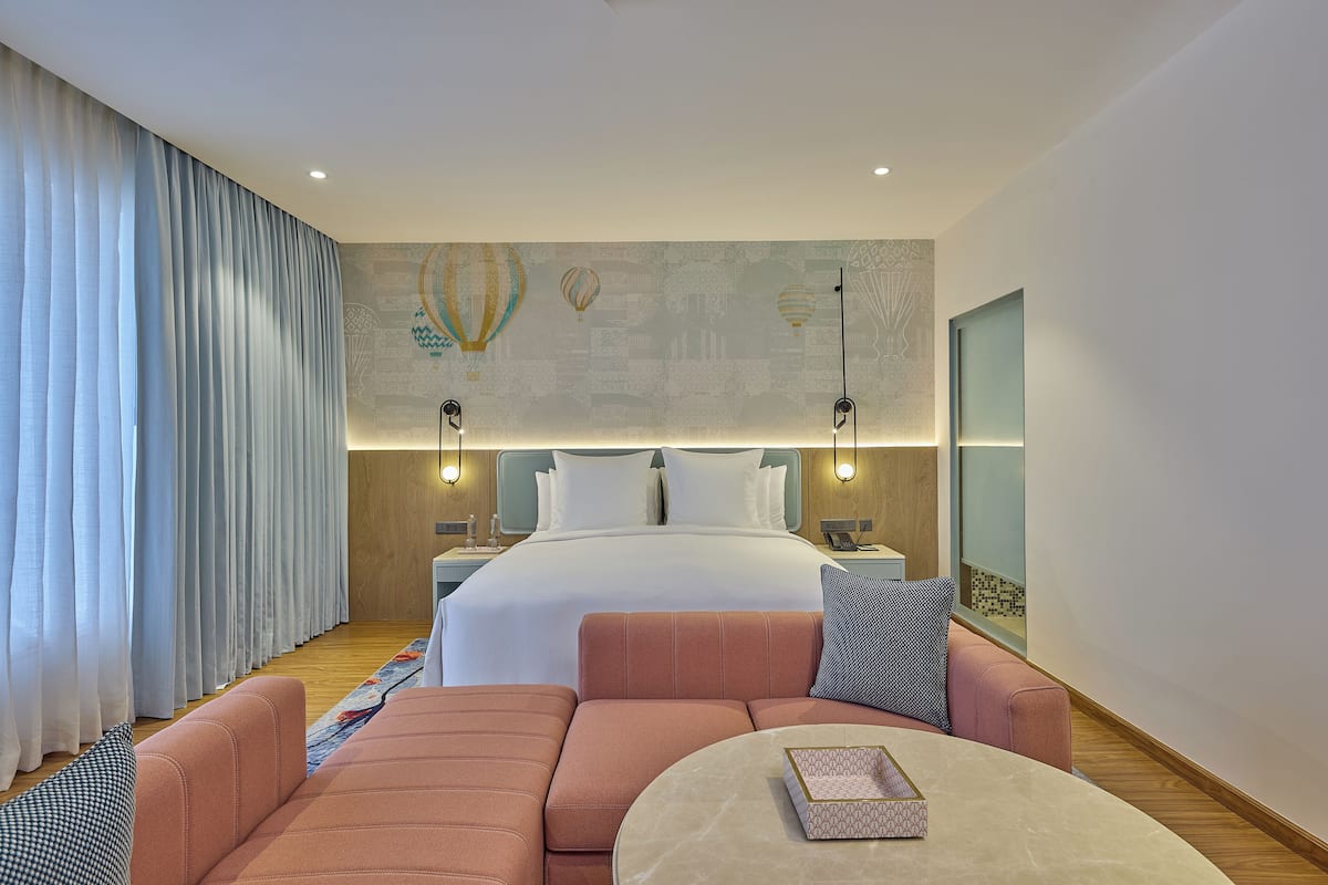 A modern hotel room with a large bed, two bedside tables, a sofa, a round coffee table, and a wall mural.