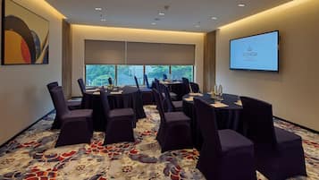 A conference room with a large screen displaying the ICONIQA HOTEL logo, a patterned carpet, and a view of a body of water through large windows.