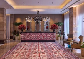 Un hall con alfombra estampada, una mesa de recepción, una araña y una zona de estar con plantas.